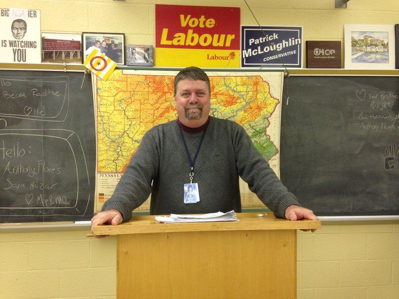 North Penn Social Studies teacher Mr. Sam Brumbaugh standing at his iconic podium in front of the map, in the famous Brumbaugh Challenge position.