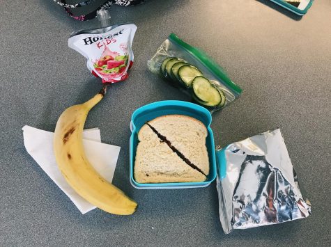 A students packed lunch consisting of a peanut butter and jelly sandwich, cucumbers, a banana, chips, and a juice box. 